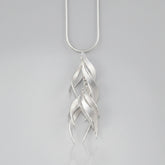A silver pendant necklace featuring a series of overlapping, stylized leaf or feather-like curved shapes hanging from a simple silver chain, set against a solid pale peach background.