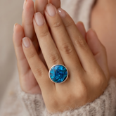 A close-up of a person's hands wearing a silver ring with a round blue marbled stone, set against a soft beige knit sweater.
