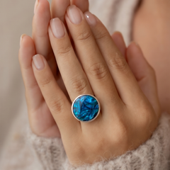A close-up of a person's hands wearing a silver ring with a round blue marbled stone, set against a soft beige knit sweater.