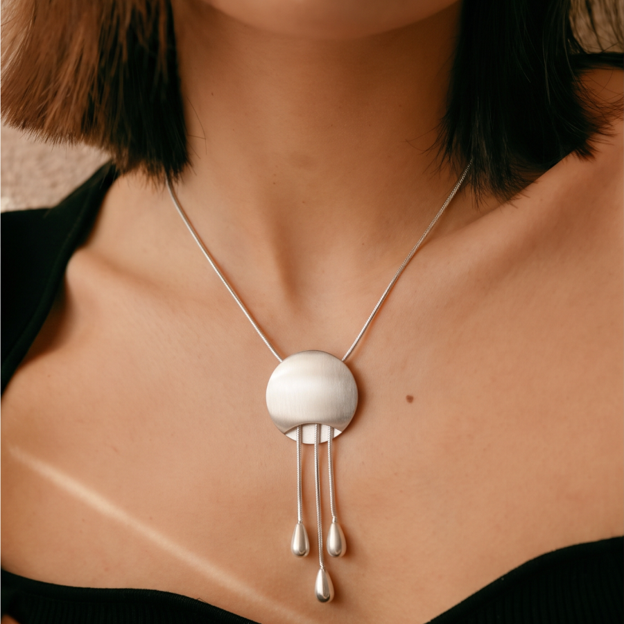 A close-up of a model wearing a silver pendant necklace with a circular disc and three dangling teardrop charms, styled with a black ribbed top.