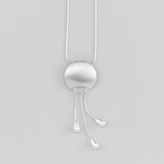 A silver pendant necklace featuring a brushed circular disc with three dangling teardrop-shaped cords, suspended against a solid background.