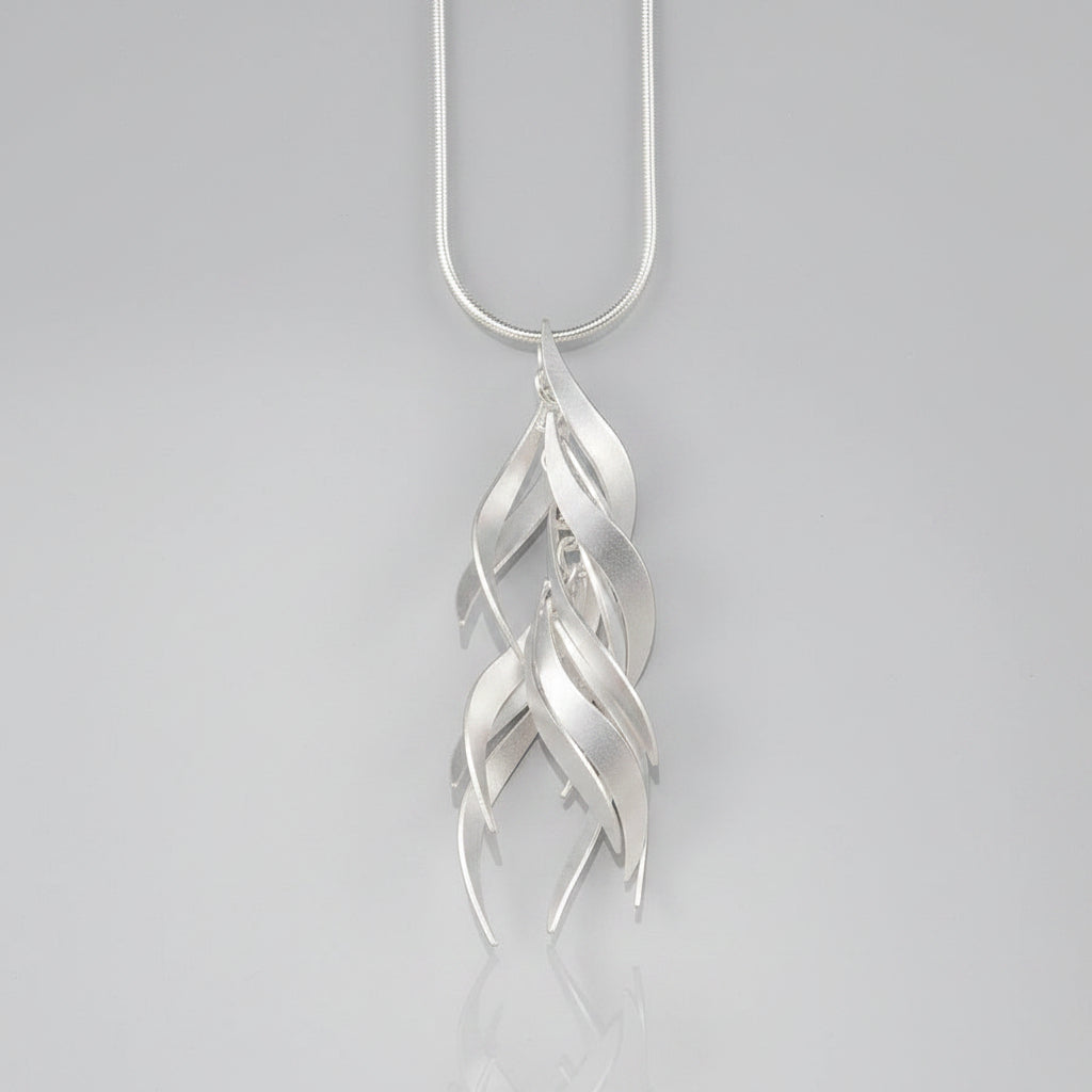 A silver pendant necklace featuring a series of overlapping, stylized leaf or feather-like curved shapes hanging from a simple silver chain, set against a solid pale peach background.