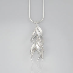 A silver pendant necklace featuring a series of overlapping, stylized leaf or feather-like curved shapes hanging from a simple silver chain, set against a solid pale peach background.