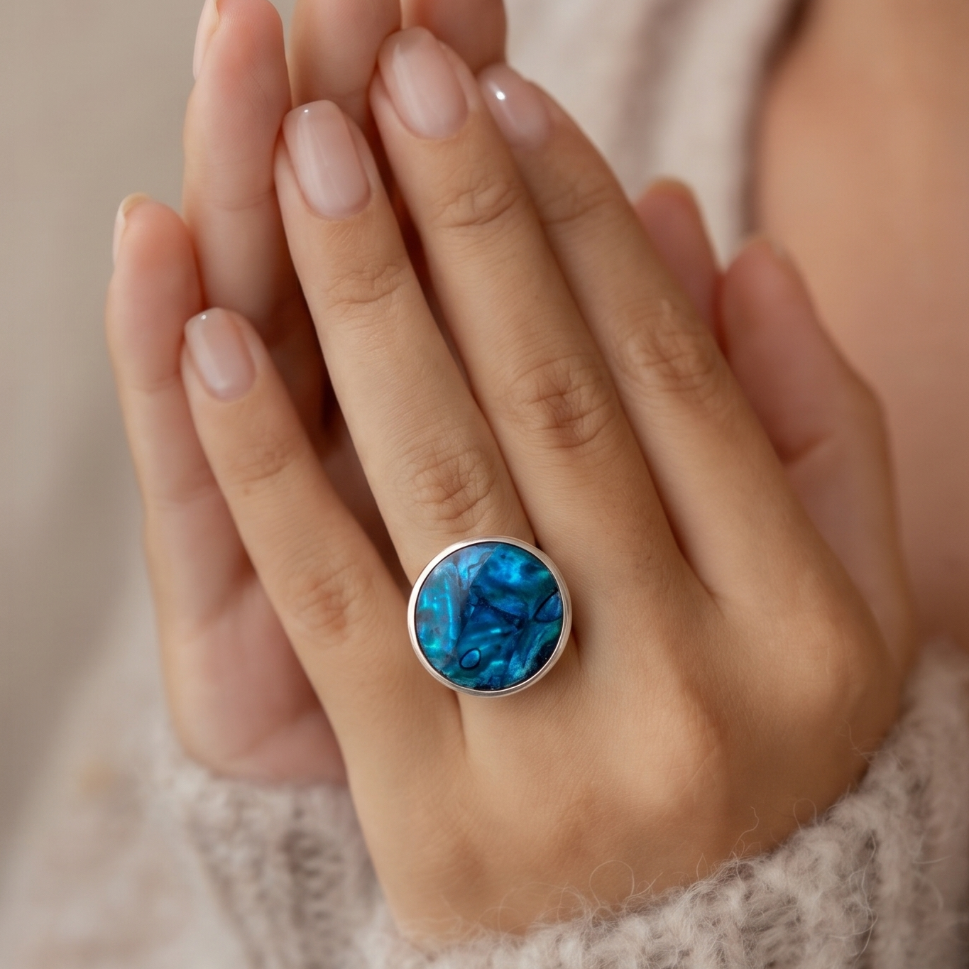 A close-up of a person's hands wearing a silver ring with a round blue marbled stone, set against a soft beige knit sweater.