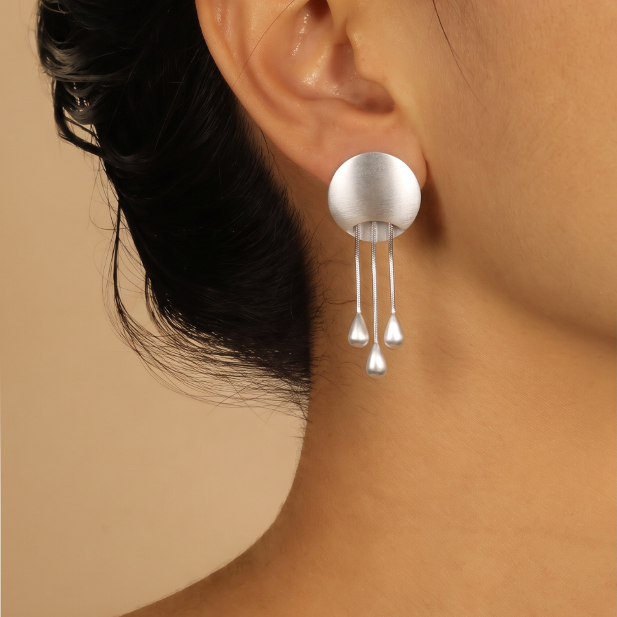 A close-up of a model's ear wearing a silver dangle earring with a matte spherical top and three hanging teardrop charms against a warm beige background.
