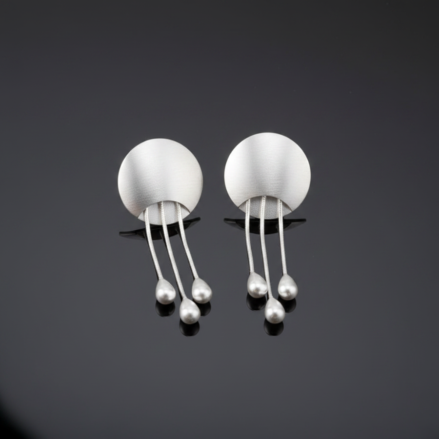 A pair of minimalist silver dangle earrings with circular disc tops and three thin vertical bars ending in small teardrop beads, displayed on a dark reflective surface.