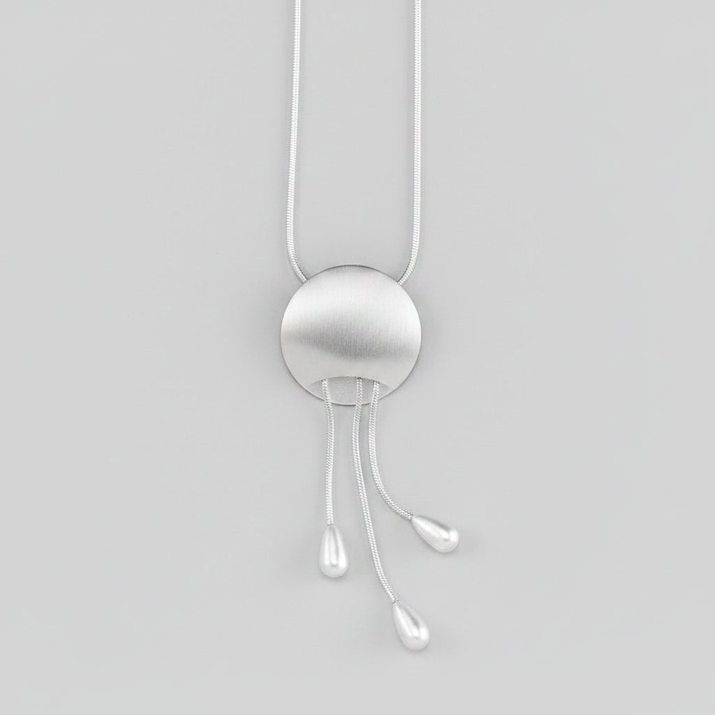 A silver pendant necklace featuring a brushed circular disc with three dangling teardrop-shaped cords, suspended against a solid background.