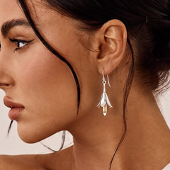 A close-up side profile of a woman wearing a delicate silver dangle earring with a floral or feather-like drop and small clear stones, against a soft light background.